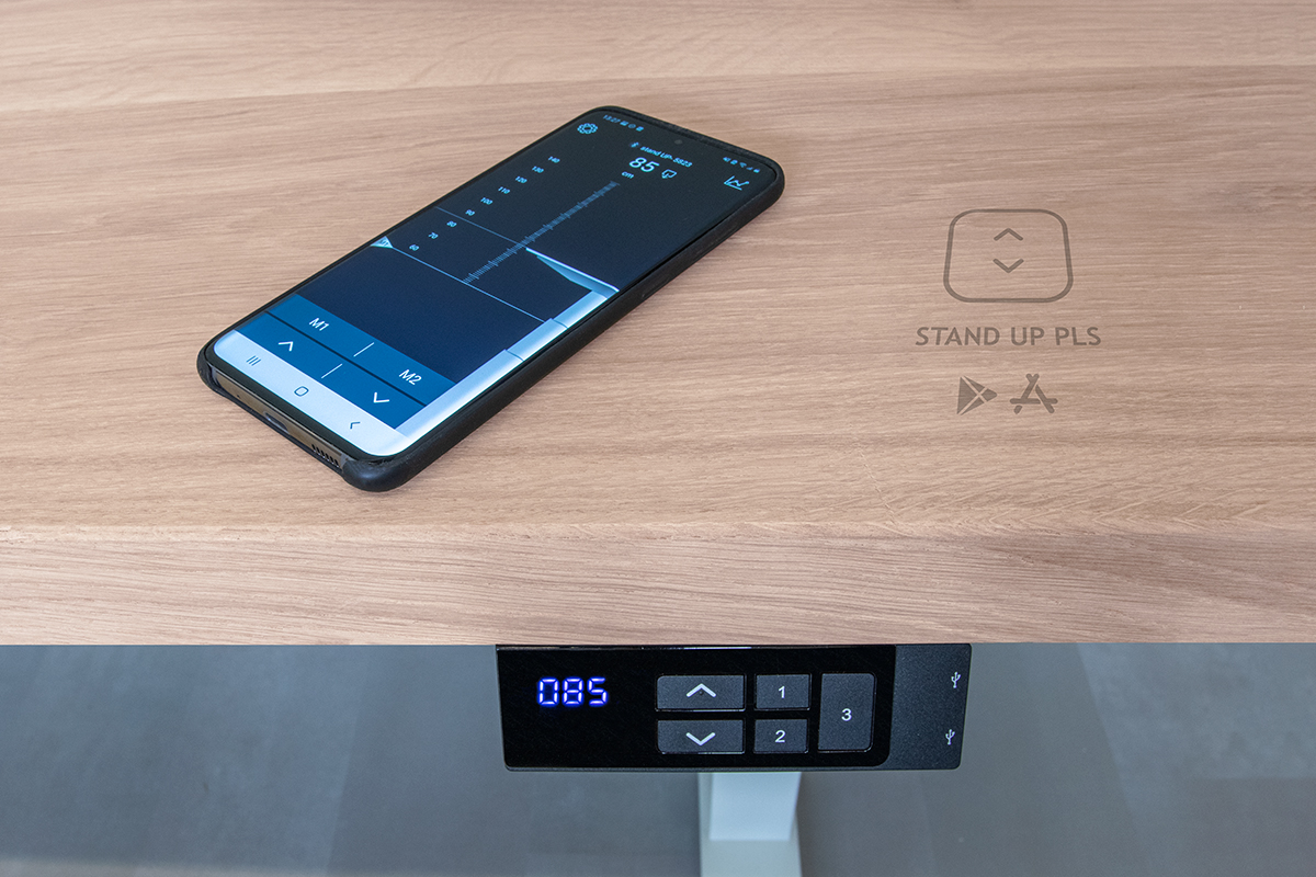 Close-up sfeerfoto van het massief eikenhouten verstelbare zit-sta bureau van GUSSTA met de mobiele stand up pls app Close-up sfeerfoto van het massief eikenhouten verstelbare zit-sta bureau van GUSSTA met de mobiele stand up pls app